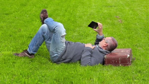 Mies matkalaukku nukkuu ruohoa ja ottaa kuvia älypuhelimella
  - Materiaali, video