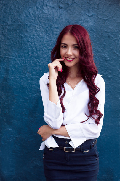 Smiling beautiful, amazing, attractive girl with long bright red hair. Glamor portrait of a sexy woman with red hair on blue background. Portrait of happy smiling red-haired, redhead girl in white blouse and blue skirt. - Фото, изображение