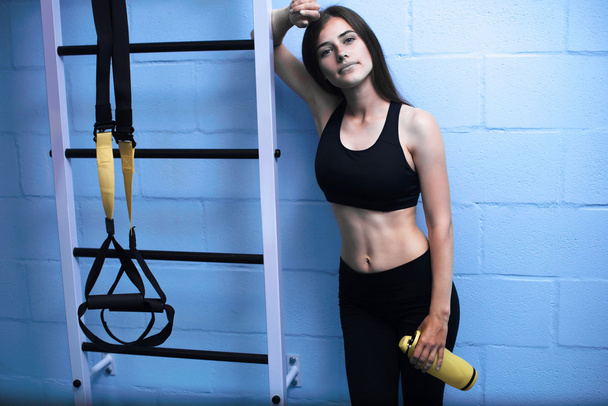 Beautiful young woman with long straight brunet hair standing with TRX near the blue wall in gym - Photo, Image