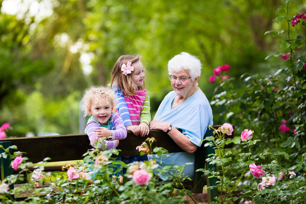 Isoäiti ja lapset istuvat ruusutarhassa
 - Valokuva, kuva