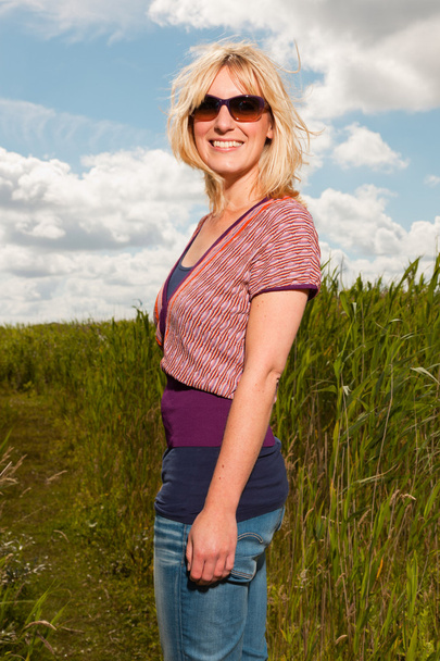 Happy pretty young blond woman with sunglasses enjoying nature. Field with high grass. Blue cloudy sky. Dressed in red. - Фото, зображення