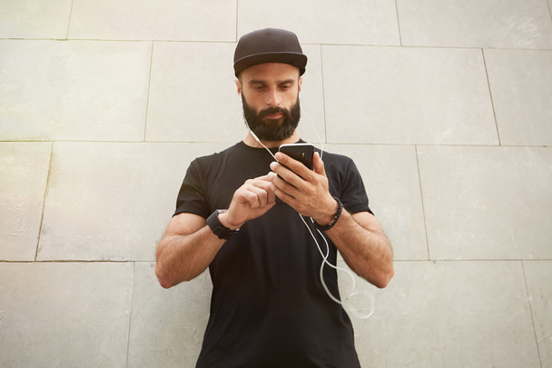 Bearded Muscular Man Wearing Black Tshirt Blank Snapback Cap Summer Time.Young Men Standing Opposite Empty Gray Concrete Wall Background Using Smartphone Headphones.Horizontal Mockup. - Photo, image