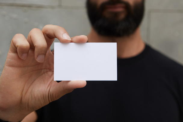 Bearded Man Wearing Casual Black Tshirt Showing Blank White Business Card.Blurred Background Ready Corporate Private Information.Horizontal Mockup. - Фото, изображение