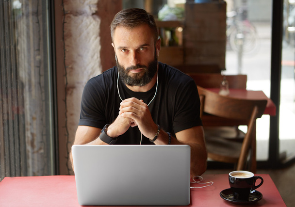 Handsome Young Bearded Businessman Wearing Black Tshirt Working Laptop Urban Cafe.Man Sitting Wood Table Cup Coffee Listening Music.Coworking Process Business Startup.Blurred Background. - Фото, изображение