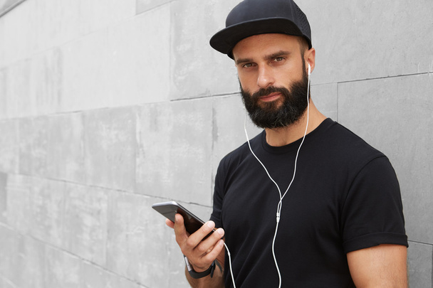 Bearded Muscular Man Wearing Black Tshirt Blank Snapback Cap Summer Time.Young Men Smiling Opposite Empty Gray Concrete Wall Background Using Smartphone Headphones.Horizontal Mockup.Color filter. - Zdjęcie, obraz