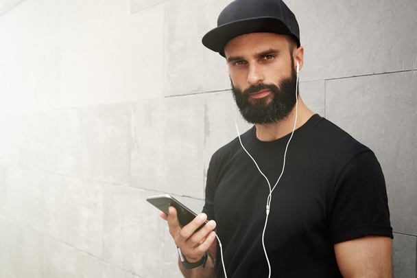 Bearded Muscular Man Wearing Black Tshirt Blank Snapback Cap Summer Time.Young Men Smiling Opposite Empty Gray Concrete Wall Background Using Smartphone Headphones.Horizontal Mockup.Color filter. - Foto, Imagen