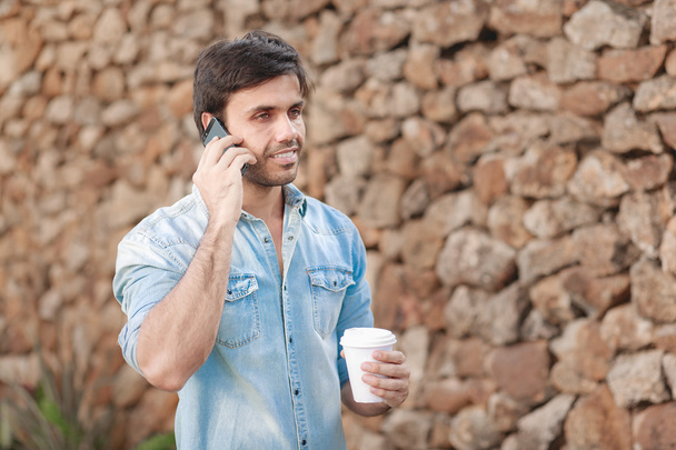 Portrait of young man drinking coffee on the street while using mobile phone - Фото, изображение