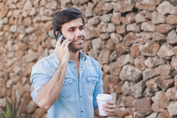 Portrait of young man drinking coffee on the street while using mobile phone - Photo, Image