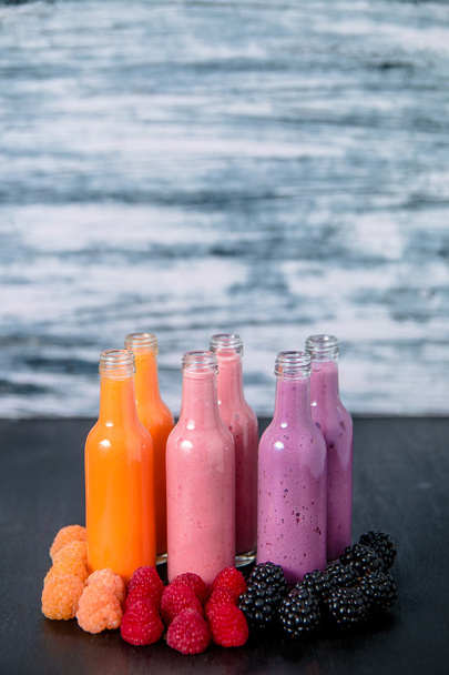 Six bottles with smoothies and raspberry, red, yellow, blackberry on black table grey background. Milk shake in glass jar berries. diet or vegan food concept, fresh vitamin - Photo, Image