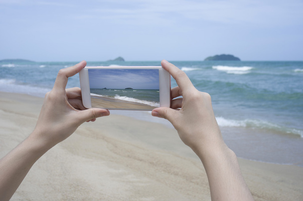 ぼやけの美しい青い海、海の写真を撮るためにスマート フォンを携帯電話、スマート フォンを持っている女性の手のクローズ アップ - 写真・画像