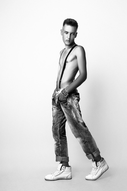 Portrait of a man with nude torso fitness standing in jeans with suspenders, on white background. Black and white - Photo, image