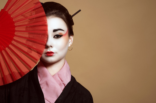 Portret van de jonge heisha in kimono verbergen van de helft van haar gezicht achter de rode handheld ventilator - Foto, afbeelding