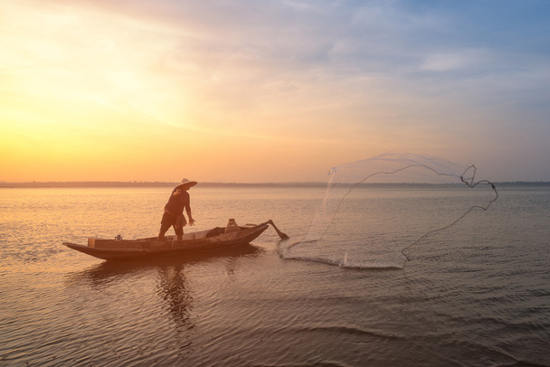 Pêcheur asiatique sur un bateau en bois jetant un filet pour attraper des poissons d'eau douce dans la rivière de la nature au début de l'heure du lever du soleil
 - Photo, image