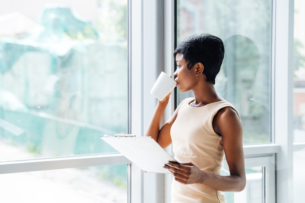 Pensive african american businesswoman with clipboard drinking coffee in office - Фото, изображение