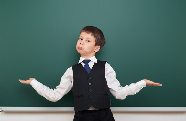 school student boy posing at the clean blackboard and open arms, grimacing and emotions, dressed in a black suit, education concept, studio photo - Photo, Image