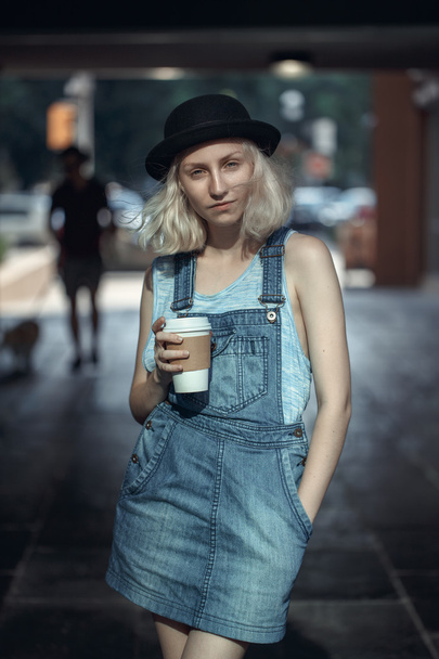 青いTシャツを着た美しい白人の若いブロンドのオルタナティブモデルの女の子の肖像画、コーヒーのカップを持つカメラで探しているジーンズロンパー、Instagramフィルターでトーン - 写真・画像