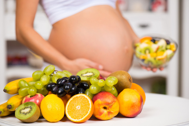 Close-up of a pregnant belly with fresh fruit and plate of salad. Healthy pregnancy, diet and vitamins - 写真・画像