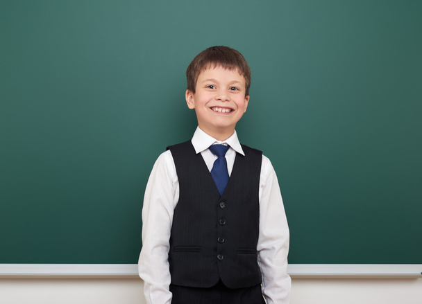 school student boy posing at the clean blackboard, grimacing and emotions, dressed in a black suit, education concept, studio photo - Photo, Image