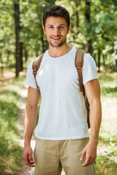 Cheerful young man with backpack standing in forest - Φωτογραφία, εικόνα