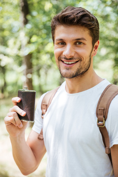 Cheerful young man standing and holding flask in forest - Fotó, kép