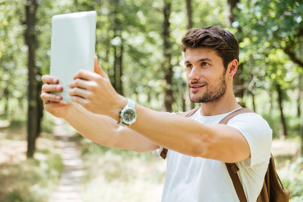 Happy man with backpack taking selfie using tablet in forest - Photo, image