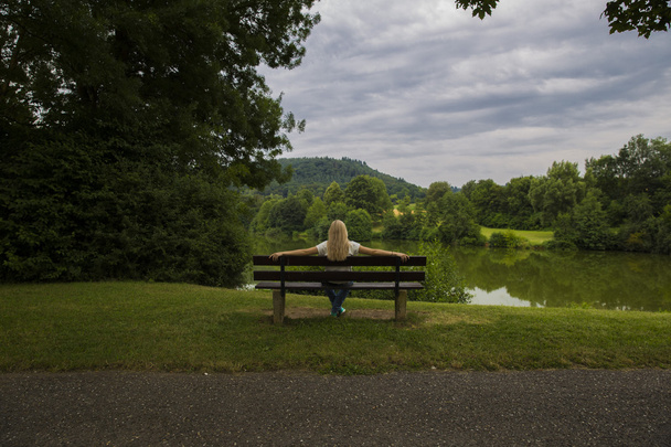 blond dívka sedí na lavičce poblíž vody a při pohledu - Fotografie, Obrázek
