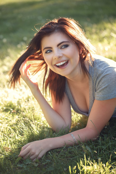 Nahaufnahme Porträt der schönen lächelnden jungen kaukasischen Frau mit rot-schwarzen Haaren, die im Freien auf Gras liegt, lachend Zähne zeigt, natürlicher Schönheit Jugendlook - Foto, Bild