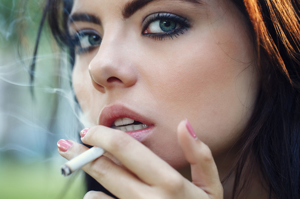 Close-up portret van witte Kaukasische mooie jonge sexy brunette vrouw met licht blauw groene ogen roken sigaret op zoek in de camera, afgezwakt met Instagram filters, slechte ongezonde gewoonte - Foto, afbeelding