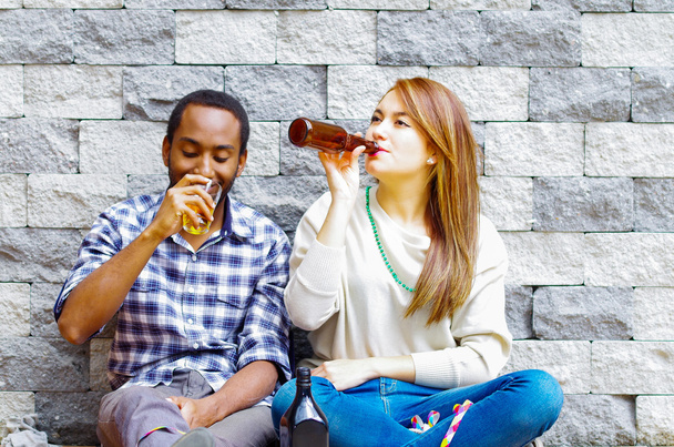 Interracial couple wearing casual clothes sitting towards grey brick wall enjoying some drinks and each others company - Foto, Bild