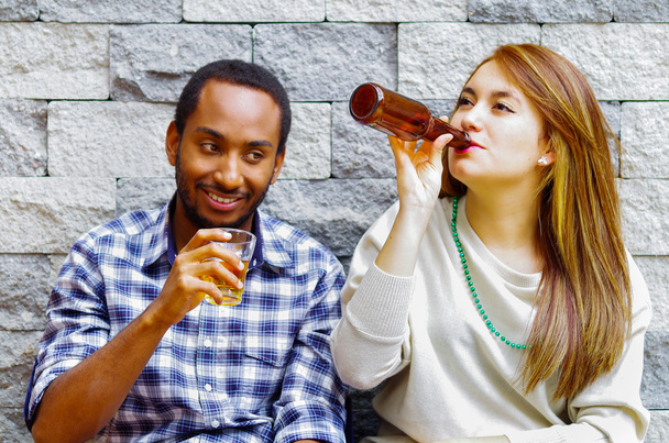 Interracial couple wearing casual clothes sitting towards grey brick wall enjoying some drinks and each others company - Φωτογραφία, εικόνα