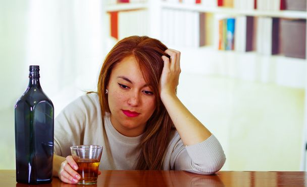 Attractive woman wearing white sweater sitting by bar counter lying over desk next to glass and bottle, drunk depressed facial expression, alcoholic concept - Photo, Image