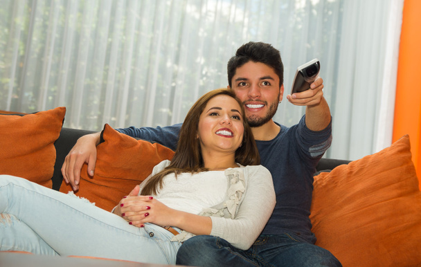 Jovem casal encantador sentado no sofá laranja abraçando e assistindo televisão, ele apontando controle remoto para câmera, ambiente albergue
 - Foto, Imagem