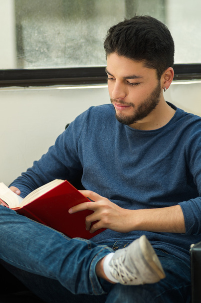 Young handsome man wearing casual clothes sitting down relaxed while reading a book, hostel guest concept - Foto, imagen