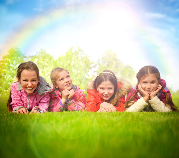 Happy group of girls lying on a green grass. Rainbow and sun ove - Foto, afbeelding
