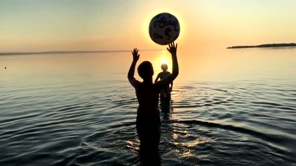Kinderen spelen bal in het water bij zonsondergang. - Video