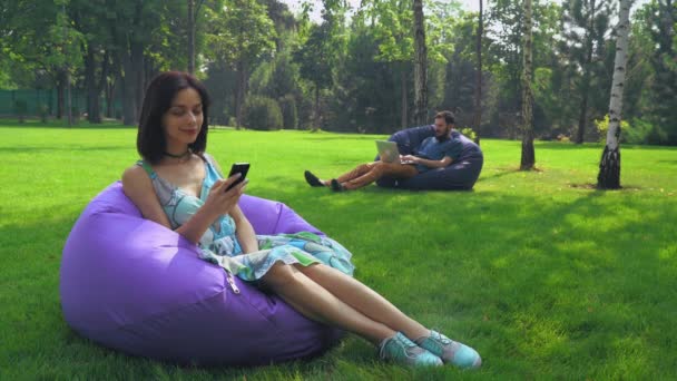 Mladá atraktivní bruneta dívka sedí v křesle v parku a získání zprávy na telefonu. - Záběry, video