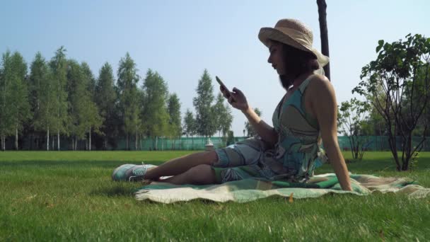 Jonge brunette meisje zittend op dekens in het park en het verkrijgen van een bericht op de telefoon. - Video
