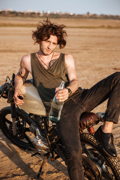 Young brutal man laying on his motorcycle and holding bottle - Photo, Image