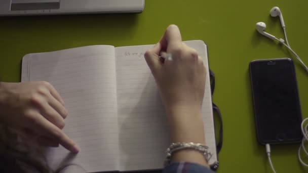Mulher mão escrevendo em notebook em branco aberto com telefone celular e fones de ouvido em uma mesa, vista superior
 - Filmagem, Vídeo