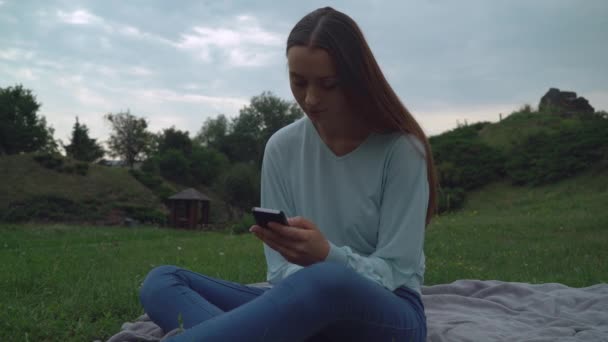Młoda dziewczyna siedzi na koce na trawnik w parku i uzyskania wiadomości w telefonie. - Materiał filmowy, wideo