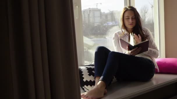 Очаровательная стильная молодая девушка читает книгу на подоконнике и смотрит в окно
 - Кадры, видео