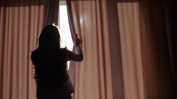 Attractive young woman opens curtains in slow motion is looking at the sunrise standing near the window in her home and enjoys of city view from height - Filmati, video