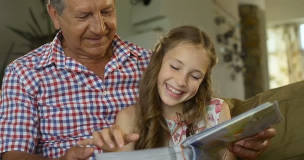 Cute girl sitting in the lap of their grandfather and looking happily together at a photo album - Footage, Video