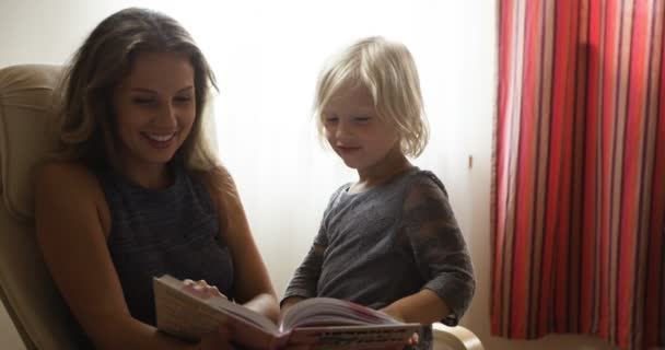 schöne blonde Mutter liest einer Tochter im Stuhl ein Buch vor - Filmmaterial, Video