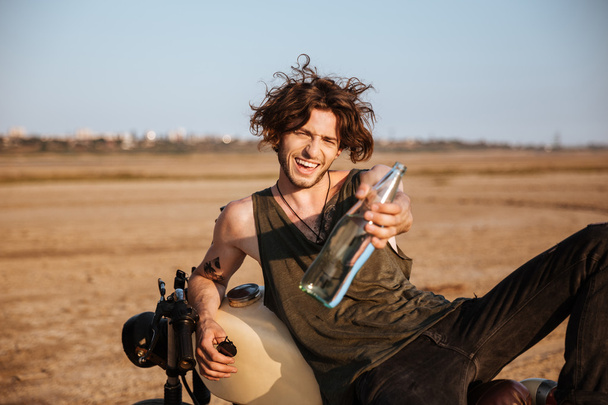 Young brutal man laying on his motorcycle and giving bottle - 写真・画像