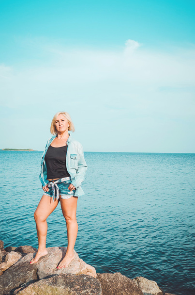 Tanned blonde mature woman walking on a beach with rock, sunny sky background, copy space. Healthy middle age female standing  coast, in denim shirt, looking at camera. Summer holiday, outdoors - Fotografie, Obrázek