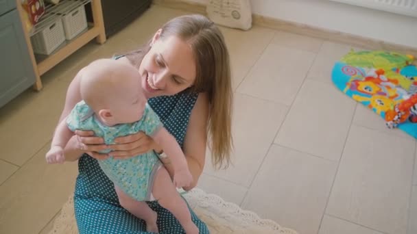 Young mother is holding her precious baby and smiling at him sitting on the kitchen floor. Slow motion - Filmagem, Vídeo