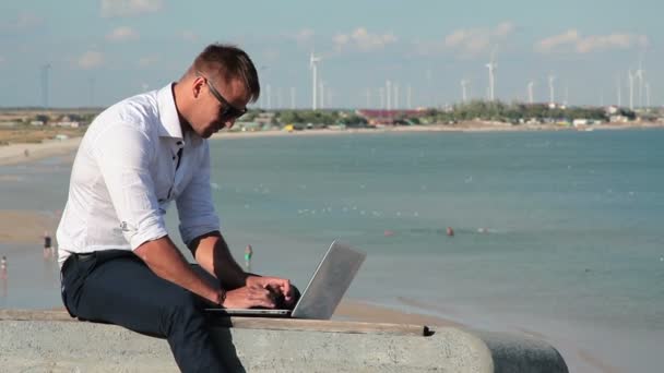 Homme d'affaires en chemise blanche et lunettes de soleil avec un ordinateur portable au bord de la mer. En arrière-plan, la mer, la rubéole, les centrales à turbine de poulet. Travailler de partout dans le monde. Freelance au travail
. - Séquence, vidéo