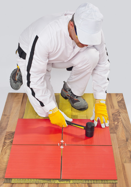 εργαζόμενος εφαρμόζει κεραμικά πλακίδια στο ξύλινο πάτωμα με ελαστικό σφυρί - Φωτογραφία, εικόνα