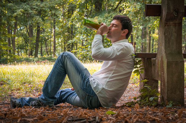 Humalainen mies makaa maassa lähellä penkkiä olutpullon kanssa
 - Valokuva, kuva